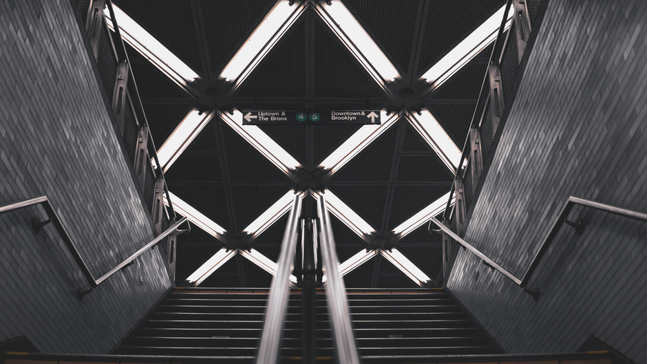 Interior view of a modern underground subway station stairway facilitating home relocation logistics, with stainless steel handrails on both sides and black metal steps leading downward. The station ceiling has a geometric pattern formed by illuminated panels with white fluorescent lighting accents. Mounted overhead signage indicates directions for uptown and The Bronx, as well as downtown and Brooklyn, referencing different subway lines, with symbols for lines 4 and 5. The surroundings include grey brick wall panels along the stairway walls, and the scene emphasizes efficient, clean passage for furniture transport and packing materials during a moving process managed by Man With a Van New Addington.