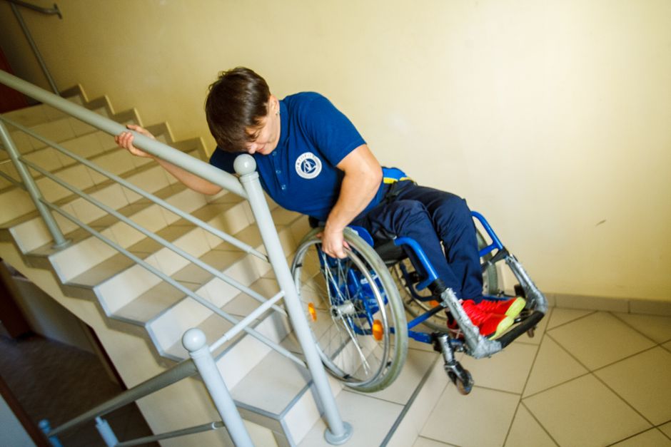 A person in a blue uniform with a logo on the chest is assisting a wheelchair user descending a staircase inside a building. The helper is holding the metal handrail with one hand while supporting the wheelchair with the other, guiding it carefully down the tiled staircase. The wheelchair is equipped with a cushioned seat, small front wheels, and larger rear wheels with orange reflectors. The individual in the wheelchair is wearing red shoes and pants, but their upper body and face are not visible. The staircase has wooden steps with a metal railing on the side, and the walls are painted in a light colour. Inside the building, the environment appears clean and well-lit. This scene demonstrates a home relocation or moving assistance process, involving careful furniture and equipment handling, which is typical of professional removals services like those provided by Man With a Van New Addington. The process highlights accessibility considerations during moving logistics, especially when navigating challenging areas such as narrow stairways or elevated entries.