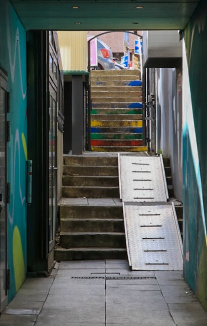 Interior view of a modern underground subway station stairway facilitating home relocation logistics, with stainless steel handrails on both sides and black metal steps leading downward. The station ceiling has a geometric pattern formed by illuminated panels with white fluorescent lighting accents. Mounted overhead signage indicates directions for uptown and The Bronx, as well as downtown and Brooklyn, referencing different subway lines, with symbols for lines 4 and 5. The surroundings include grey brick wall panels along the stairway walls, and the scene emphasizes efficient, clean passage for furniture transport and packing materials during a moving process managed by Man With a Van New Addington.