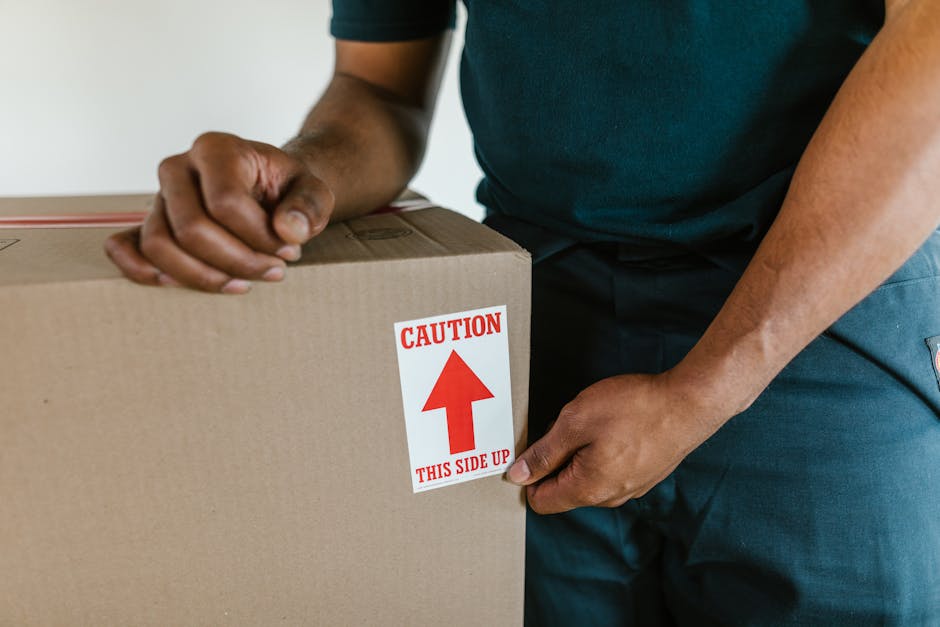A close-up image showing a person wearing a dark blue t-shirt and pants, holding a cardboard box equipped with a red and white label that reads 'CAUTION THIS SIDE UP' with a large upward-pointing arrow. The person is gripping the box firmly with their right hand at the bottom and their left hand placed on top, indicating careful handling during a home relocation or furniture transport process. The box is positioned inside what appears to be a doorway or entrance, with part of their body visible, standing on a light-colored floor. The background is blurred but suggests an indoor environment, possibly the interior of a house or flat. This image exemplifies proper packing and handling procedures, which are key aspects of professional removals services offered by Man With a Van New Addington, demonstrating careful loading techniques during packing and moving operations.