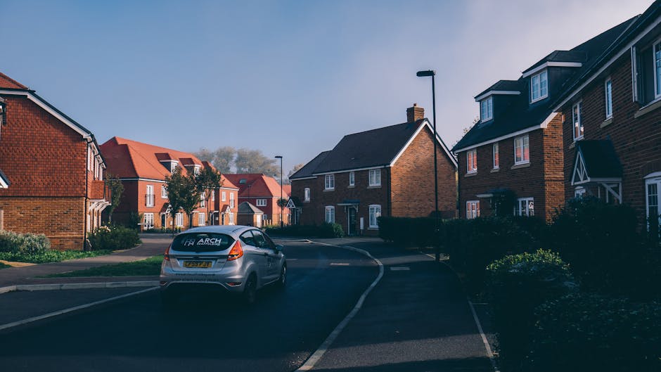 A residential street scene showing a silver van parked on the pavement adjacent to a modern brick house with a driveway. The van, branded with 'Man With a Van New Addington,' is positioned near the entrance of a home relocation or furniture transport operation, with the rear doors open and the vehicle partially loaded. Several cardboard boxes, wrapped furniture, and packing materials such as plastic wrap and blankets are visible inside or being carried towards the van. The street is lined with similar detached and semi-detached houses featuring red or dark roofing, with some having small front gardens and hedges. Streetlights are mounted on poles along the road, and the scene is illuminated by daylight under a clear sky, suggesting an outdoor loading process within a typical suburban neighbourhood. This setup reflects the logistics involved in professional removals and packing services, supporting house moves in Addington Village.
