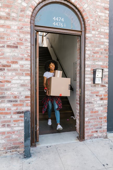 A close-up image showing a person wearing a dark blue t-shirt and pants, holding a cardboard box equipped with a red and white label that reads 'CAUTION THIS SIDE UP' with a large upward-pointing arrow. The person is gripping the box firmly with their right hand at the bottom and their left hand placed on top, indicating careful handling during a home relocation or furniture transport process. The box is positioned inside what appears to be a doorway or entrance, with part of their body visible, standing on a light-colored floor. The background is blurred but suggests an indoor environment, possibly the interior of a house or flat. This image exemplifies proper packing and handling procedures, which are key aspects of professional removals services offered by Man With a Van New Addington, demonstrating careful loading techniques during packing and moving operations.
