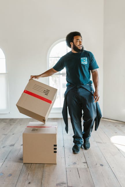 A man with a beard, wearing a navy blue t-shirt and dark trousers, is standing inside a bright, empty room with light-colored wooden flooring and a large arched window in the background. He is holding a cardboard box with red and black markings, preparing to place it on the floor or onto another box. The room appears to be part of a home undergoing a packing and moving process, with minimal furnishings or decorations visible. The natural light coming through the window illuminates the space, highlighting the clean and open environment suitable for house removals. Man With a Van New Addington, a professional removals company, specializes in home relocation and furniture transport, supporting clients through efficient packing and loading procedures. The scene captures the logistics of loading boxes and packing materials, emphasizing careful handling during the home relocation process.