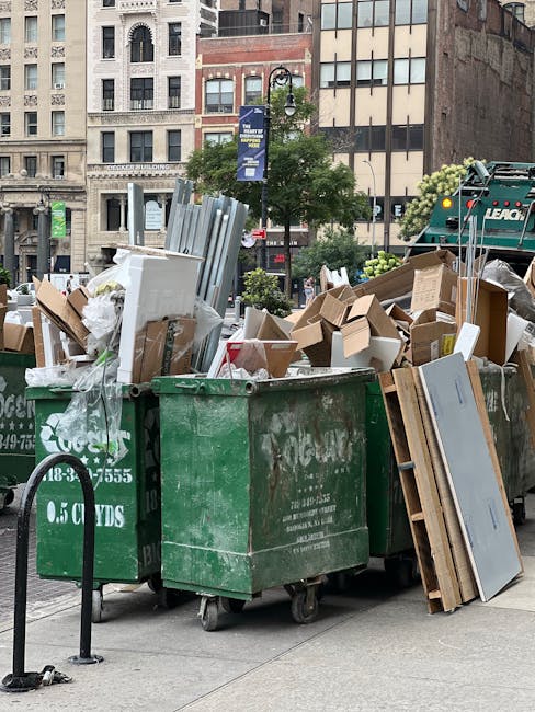 A row of three large green waste bins filled with discarded household items, including flattened cardboard boxes, plastic packaging, and pieces of wood and metal, situated on a city pavement in front of multi-story buildings with various window styles and brick facades. Some boxes and materials are protruding from the bins, with a metal trolley with straps visible nearby, indicating collection for waste or recycling. The scene is outdoors during daytime with natural light illuminating the urban environment. This setup may relate to house removals or clearing services provided by Man With a Van New Addington, illustrating the typical disposal process during a home relocation or clearance.