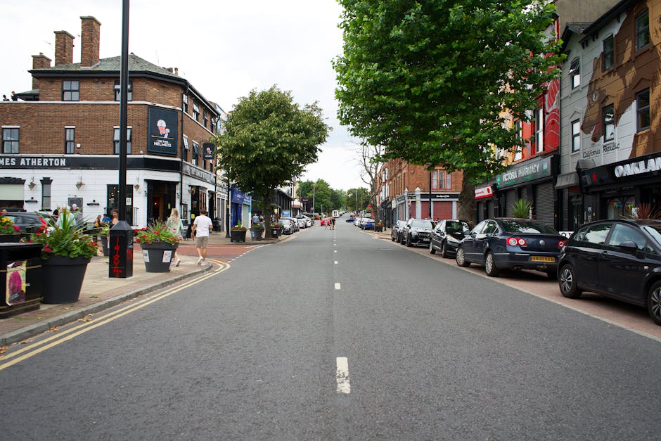 A quiet urban street scene on a cloudy day with a clear, straight road running through the center, lined on both sides by parked cars including sedans and hatchbacks, with some vehicles partially visible. On the left side, there is a sidewalk with large planter pots filled with flowers and green foliage, a few pedestrians walking past the storefronts of cafes and shops, and a lamppost near a building with signage for local businesses such as a pub or restaurant. On the right, more shops with colorful facades and awnings are visible, some with signs for pharmacy and retail outlets. In the background, trees with green leaves line the street, providing shade and a touch of nature in the urban environment. This setting illustrates a typical scene where a house removal or furniture transport process may be taking place, with Man With a Van New Addington and their home relocation services potentially involved in maneuvering moving equipment or vehicles nearby in this familiar town centre area.