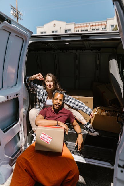 Two women are inside the open rear of a cargo van parked outdoors on a clear day, surrounded by residential buildings and utility poles. One woman, seated on the van's edge, is wearing a maroon top and orange skirt, holding a cardboard box labeled 'FRAGILE,' and smiling at the camera. The second woman, standing inside the van, is wearing a black and white checkered shirt over a white top, with her arm resting on the doorframe, also smiling. The van's interior contains several cardboard boxes of various sizes, some stacked, and packing materials such as plastic wraps and paper. The scene depicts a home relocation and furniture transport process, highlighting packing and loading activities as part of house removals services by Man With a Van New Addington.