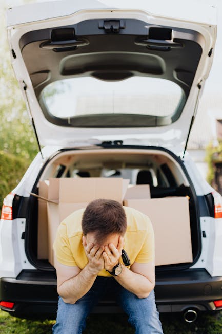 A man wearing a yellow T-shirt and blue jeans is crouching in front of an open white hatchback vehicle, holding his face in his hands with a distressed expression. The car's trunk is filled with large cardboard boxes and packing materials, indicating an ongoing home removal or furniture transport process. The vehicle is parked outdoors on a grassy area, with a background that includes trees and a partly cloudy sky. Visible inside the vehicle are additional boxes and packing items, suggesting that the man is involved in packing or organizing belongings for a house move, with the scene representing the logistics of packing and loading for a local relocation service provided by Man With a Van New Addington.