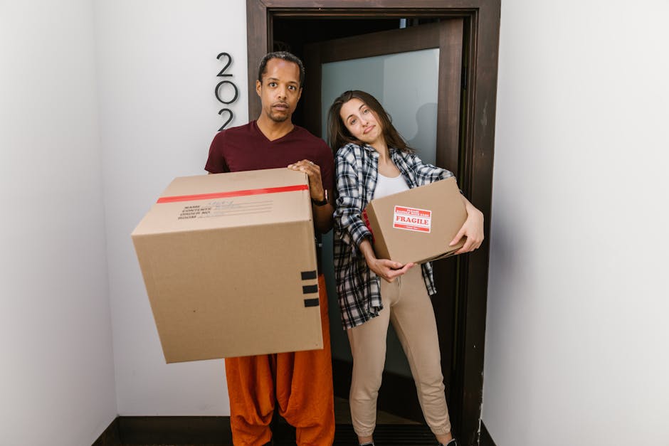 Two women are inside the open rear of a cargo van parked outdoors on a clear day, surrounded by residential buildings and utility poles. One woman, seated on the van's edge, is wearing a maroon top and orange skirt, holding a cardboard box labeled 'FRAGILE,' and smiling at the camera. The second woman, standing inside the van, is wearing a black and white checkered shirt over a white top, with her arm resting on the doorframe, also smiling. The van's interior contains several cardboard boxes of various sizes, some stacked, and packing materials such as plastic wraps and paper. The scene depicts a home relocation and furniture transport process, highlighting packing and loading activities as part of house removals services by Man With a Van New Addington.