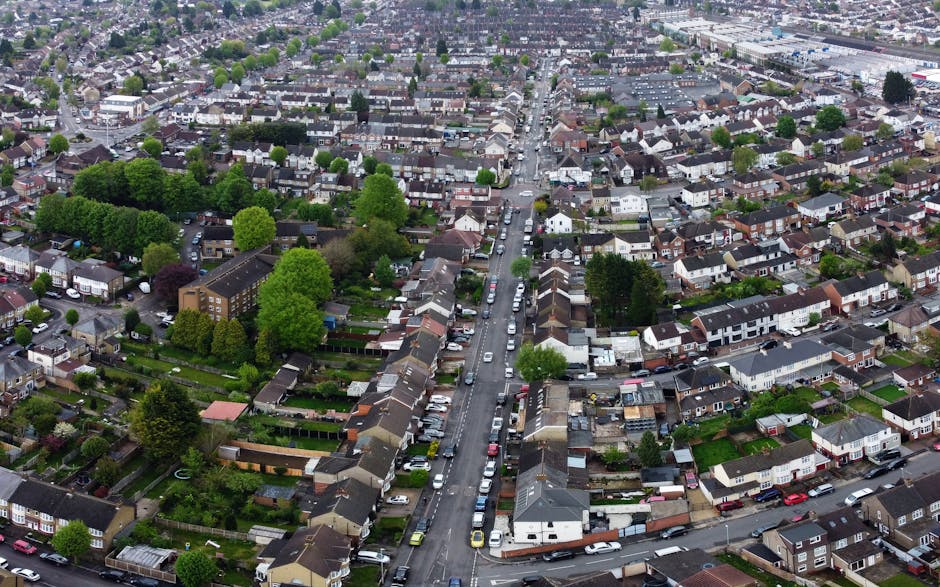 An aerial view of a residential neighbourhood showing a wide street lined with parked cars on both sides, flanked by rows of terraced houses with sloped roofs. There are numerous mature trees interspersed among the houses, providing greenery throughout the area. In the foreground, a section of a private driveway with additional parked vehicles leads to back gardens, some with sheds or small structures. The image captures a calm, well-organized urban environment typical of a suburban home relocation area, with no visible moving equipment or personnel present, emphasizing the residential setting suitable for house removals and furniture transport services provided by companies like Man With a Van New Addington.