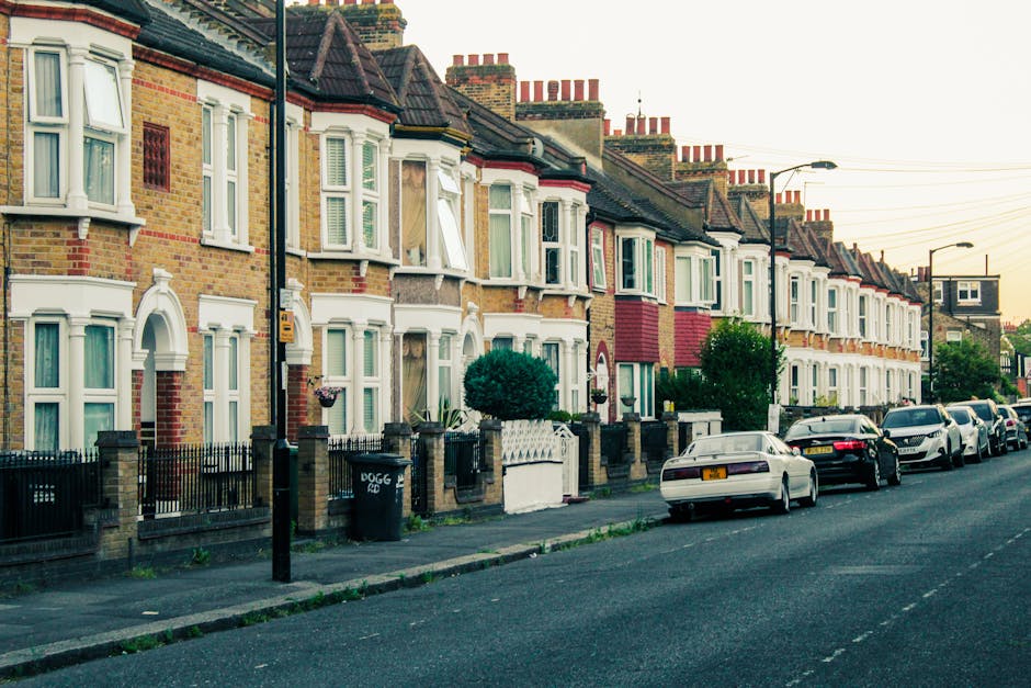 An aerial view of a residential neighbourhood showing a wide street lined with parked cars on both sides, flanked by rows of terraced houses with sloped roofs. There are numerous mature trees interspersed among the houses, providing greenery throughout the area. In the foreground, a section of a private driveway with additional parked vehicles leads to back gardens, some with sheds or small structures. The image captures a calm, well-organized urban environment typical of a suburban home relocation area, with no visible moving equipment or personnel present, emphasizing the residential setting suitable for house removals and furniture transport services provided by companies like Man With a Van New Addington.