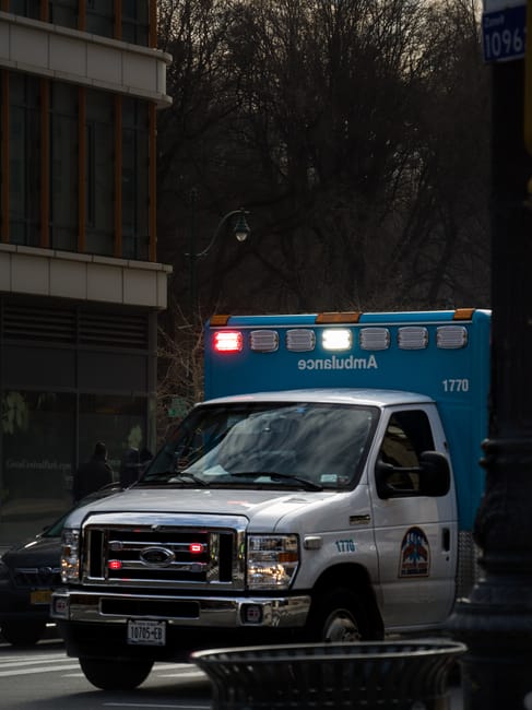 A white van with blue emergency lights on top, marked with the number 1770 and the logo of an ambulance service, is parked on a city street in New Addington. The van is positioned near a building with a large glass window and is part of a home relocation or furniture transport operation, as part of the loading process for house removals. Several cardboard boxes and household items are visible inside the vehicle through the windshield, indicating preparations for moving or packing. The area appears to be a busy urban environment with trees in the background and streetlamps, suggesting it may be late afternoon or early evening. Man With a Van New Addington’s services are associated with efficient and professional removals, often involving careful loading and transportation of furniture, boxes, and appliances, as seen in this scene of a vehicle equipped for emergency or urgent moves by a professional removal company.