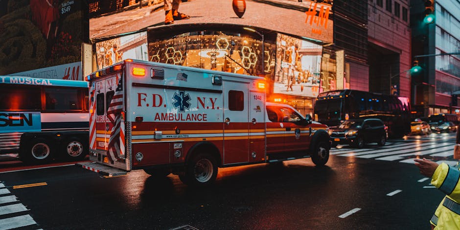 A red and white FDNY ambulance is positioned on a city street during the evening, with its emergency lights flashing. The ambulance is centrally located in the image, slightly to the left, and is surrounded by other vehicles including a black van and a bus, both parked along the curb. The street is wet, reflecting the vehicle lights, and there is a pedestrian wearing a bright yellow jacket visible on the right side, partially cropped, possibly directing or observing the scene. In the background, a large modern building features illuminated advertising screens and storefront windows, with some displays showing bright, colorful patterns. The scene appears to be in an urban area with dense traffic and illuminated commercial signage, possibly during a busy evening or night-time. Man With a Van New Addington's services relate to house removals and moving logistics, which might be relevant in terms of city environment imagery, although this specific photo depicts an emergency vehicle during a response, emphasizing urgent transportation and city logistics.