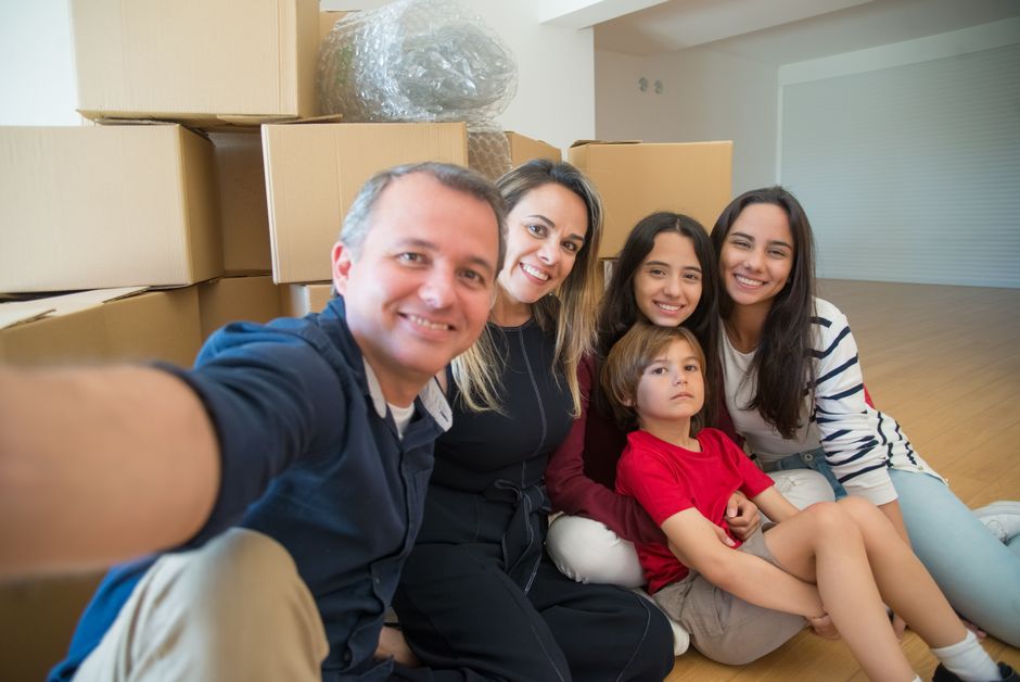 A family of five, including two adults and three children, sitting on the wooden floor of a packed living room surrounded by numerous cardboard boxes, plastic-wrapped furniture, and household items, i