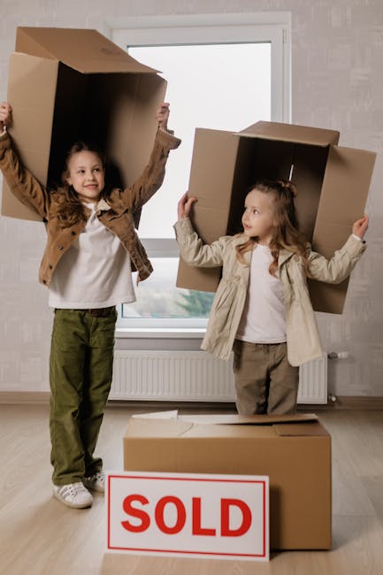 A family of five, including two adults and three children, sitting on the wooden floor of a packed living room surrounded by numerous cardboard boxes, plastic-wrapped furniture, and household items, i