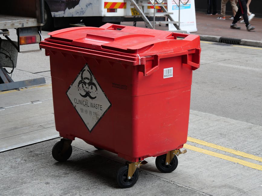 A red clinical waste bin with a hinged lid, mounted on four black caster wheels, positioned on a concrete pavement near the entrance of a property during a home relocation process. The bin displays a hazardous biohazard symbol and a 'Clinical Waste' warning label. In the background, part of a white vehicle or removal truck is visible, with a metal staircase or ramp leading up to the vehicle, indicating loading or transport activity. Surrounding the scene are scattered pedestrians and street elements, including yellow parking restriction lines on the road. The setting suggests it is part of the packing and moving logistics managed by Man With a Van New Addington, with a focus on safe disposal of hazardous items during a house removal in New Addington.