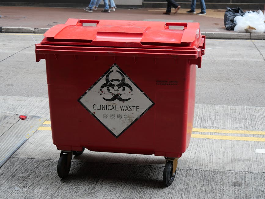 A yellow metal needle drop box with a biohazard symbol and a padlock at the base, positioned on a hard surface against a dark grey wall. The box has a slot labeled 'Needle Drop Box' and is marked with stickers, including one with the biohazard icon and another for secure disposal. The surrounding area includes a partially visible graffiti-covered surface on the left and scattered debris at the bottom, suggesting an urban environment. This container is used for disposing of hazardous waste, supporting safe disposal laws during home relocations or moving processes conducted by Man With a Van New Addington, especially when handling items requiring specialised disposal.
