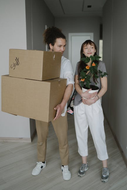 A woman with curly hair, dressed in beige trousers and a white top, is standing inside a modern, well-lit hallway holding two cardboard boxes, one stacked on top of the other, with handwritten labels on the sides. She appears to be preparing for a home relocation, with the boxes likely containing household items. Next to her, a young girl with straight hair, wearing a grey top, white pants, and grey sneakers, is holding a potted houseplant with green foliage and orange flowers, possibly moving it to a new residence. The hallway has plain grey walls and a laminate floor, and a doorway is visible behind them. The scene depicts the packing and moving process typical of furniture transport and home packing activities. Occasionally, Man With a Van New Addington's removals services are associated with this type of relocation work, including loading, packing, and transport of household belongings.