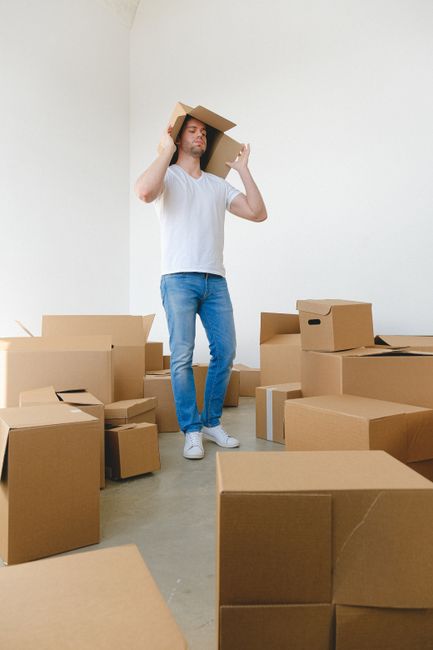 A man with light skin, wearing a white t-shirt, blue jeans, and white sneakers, is standing inside a room with white walls and a concrete floor, surrounded by numerous cardboard boxes of various sizes, some open and some sealed with packing tape. He is holding a medium-sized cardboard box on his shoulder and appears to be in the process of packing or preparing for a home relocation. The boxes are arranged around him, some stacked and others scattered on the ground, indicating an active moving or packing process. The room is well-lit with natural light, suggesting a daytime setting. This scene is reflective of furniture transport and packing activities associated with house removals, and it showcases the typical steps involved in furniture transport and loading during a clean, organized move. The company Man With a Van New Addington provides removals services that cover these aspects of home relocations.
