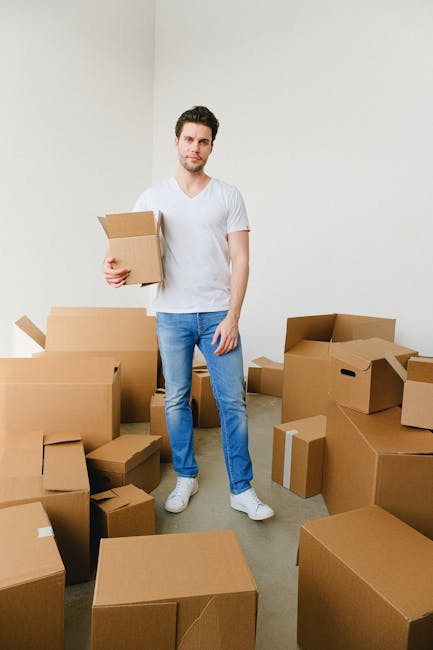 A woman with curly hair, dressed in beige trousers and a white top, is standing inside a modern, well-lit hallway holding two cardboard boxes, one stacked on top of the other, with handwritten labels on the sides. She appears to be preparing for a home relocation, with the boxes likely containing household items. Next to her, a young girl with straight hair, wearing a grey top, white pants, and grey sneakers, is holding a potted houseplant with green foliage and orange flowers, possibly moving it to a new residence. The hallway has plain grey walls and a laminate floor, and a doorway is visible behind them. The scene depicts the packing and moving process typical of furniture transport and home packing activities. Occasionally, Man With a Van New Addington's removals services are associated with this type of relocation work, including loading, packing, and transport of household belongings.