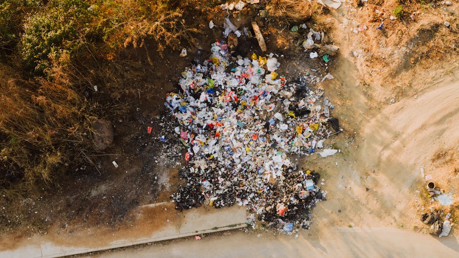 An aerial view of a large pile of mixed household waste and rubbish dumped on a patch of bare ground beside a paved sidewalk, with some vegetation and shrubs nearby. The trash includes crumpled plastic containers, cardboard boxes, plastic bags, paper debris, and small fragments of various materials. The surrounding area shows exposed earth and dirt, with a concrete curb at the bottom edge of the image. No vehicles or people are visible in this scene. The image appears to depict illegal dumping or waste disposal near a residential or urban area, highlighting the importance of proper waste management and adherence to local dumping rules to avoid fines, as outlined in the 'Skip and Dumping Rules in New Addington' webpage by Man With a Van New Addington.