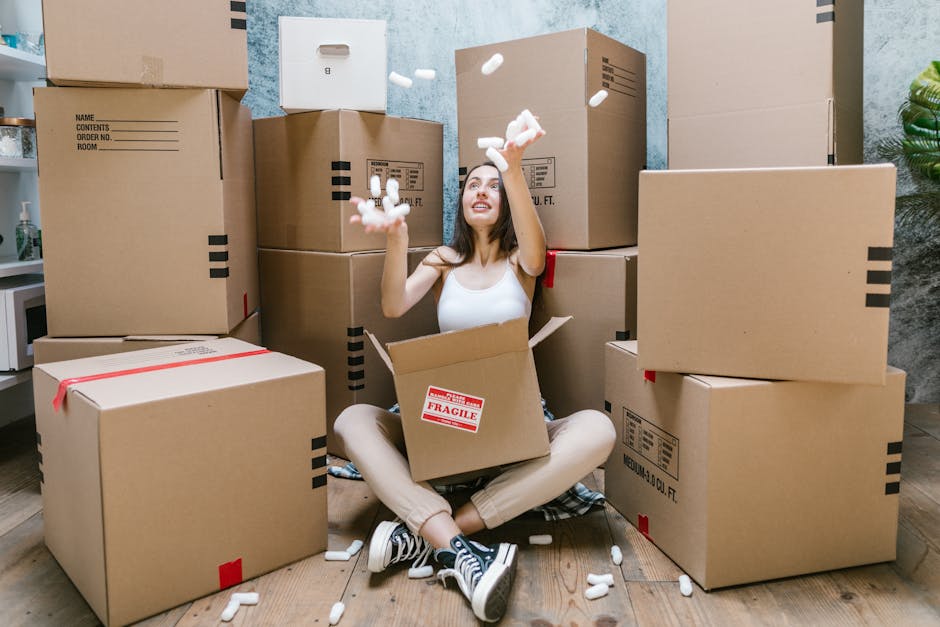 A young woman with long dark hair, sitting cross-legged on a wooden floor inside a property, is surrounded by a variety of large cardboard moving boxes, some labeled with property or content details. She is in the process of packing or unpacking, with her arms raised and a cheerful expression, as she tosses white packing peanuts into the air. The boxes are stacked on all sides of her, with some placed on the floor and others arranged against a textured blue wall. A cardboard box with a red 'Fragile' sticker is positioned on her lap. To her left, a small white shelf is partially visible, with a potted plant nearby. The scene depicts a typical home relocation or packing process, with the setup indicating careful preparation for a furniture transport or house removal service offered by Man With a Van New Addington, focusing on efficient moving logistics and packing practices.